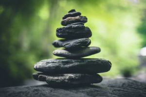 Una serie di pietre scure e piatte messe una sull'altra a creare una piccola piramide irregolare che resta in equilibrio con uno delle foglie verdi sullo sfondo