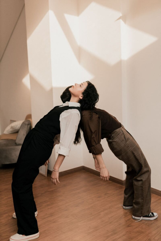 Due persone in posa artistica, schiena contro schiena e piegate all’indietro a formare un arco, in una stanza con pareti chiare e pavimento in legno; indossano abiti eleganti casual, mentre sulla parete si proiettano forme geometriche di luce.
